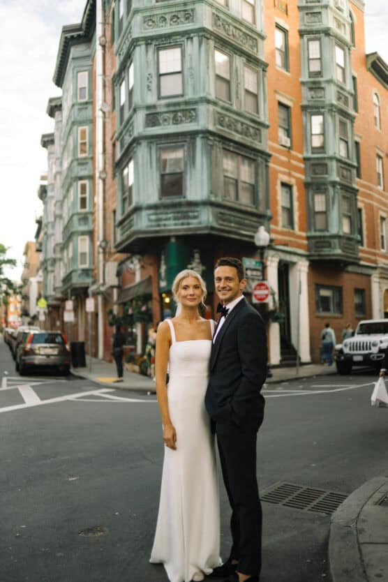 A bride in a white dress and a groom in a black tuxedo stand together, smiling, on a city street corner with a historic building featuring green bay windows in the background. by Justin Salem Meyer