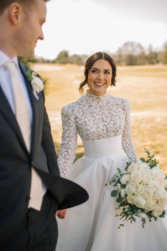 A bride in a long-sleeved lace wedding dress holds a bouquet of white roses and greenery, smiling at the groom in a dark suit and white tie as they walk outdoors on a sunny day. by Justin Salem Meyer