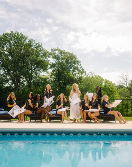Ten women sit on lounge chairs by a pool, some reading newspapers, while one stands in the center. They are outdoors, surrounded by trees, with a bright sky above. by Justin Salem Meyer