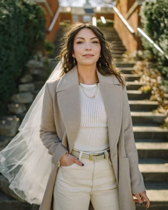 A woman in a beige coat and white outfit stands outdoors on stone steps, wearing a veil. She has long wavy hair and is looking at the camera, with one hand in her pocket. The background shows greenery and brick walls. by Justin Salem Meyer