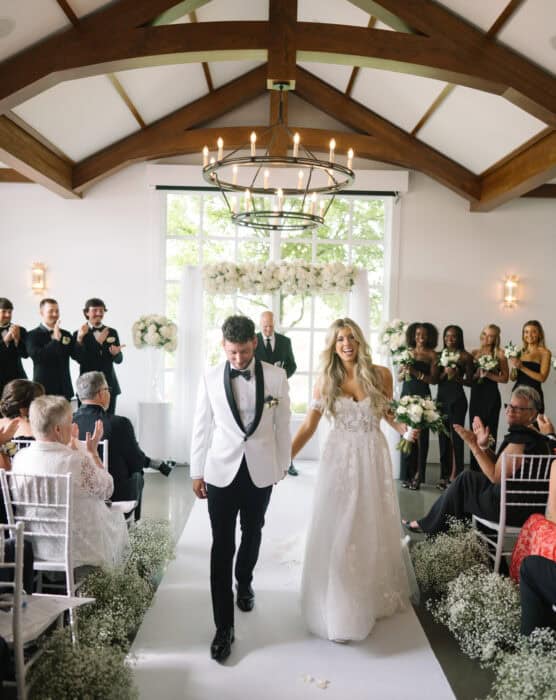 A newlywed couple walks down the aisle holding hands, smiling, surrounded by clapping guests in a bright, elegant venue with white flowers and wooden beams overhead. by Justin Salem Meyer