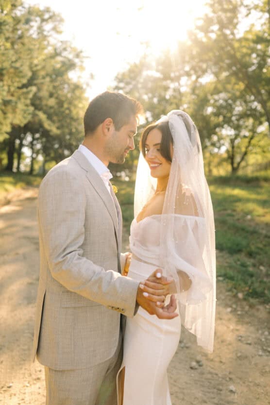 A bride and groom stand outdoors on a sunlit path, smiling and holding hands. The bride wears a white dress and veil, while the groom wears a light beige suit. Trees and sunlight create a warm, romantic atmosphere. by Justin Salem Meyer