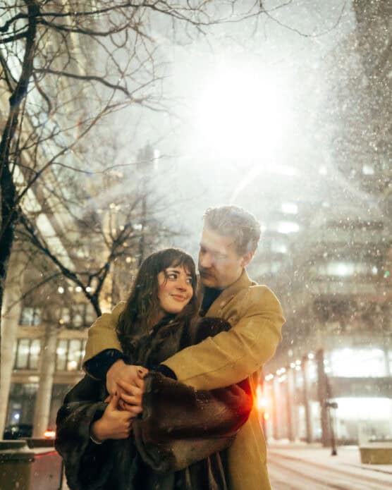 A couple embraces warmly on a snowy city street at night, illuminated by bright streetlights, with bare trees and tall buildings in the background. by Justin Salem Meyer
