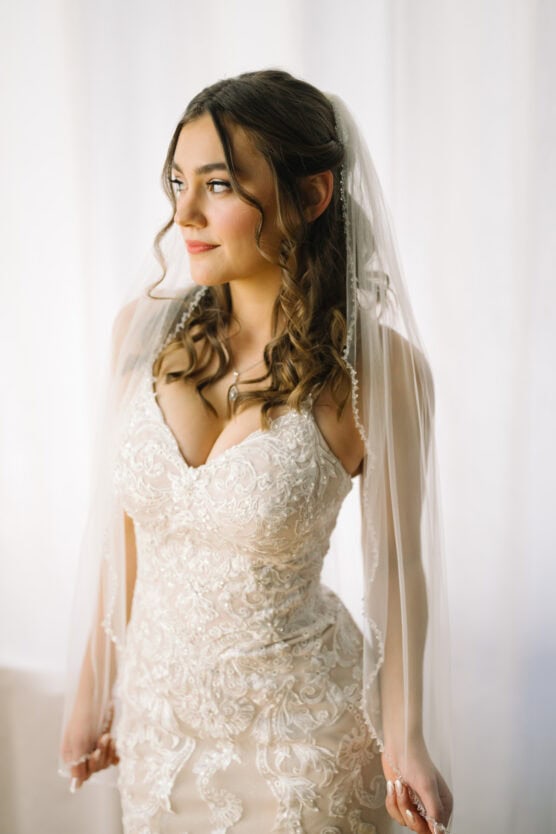 A bride with wavy brown hair in a white lace wedding dress and veil stands in soft lighting, looking to the side with a gentle expression. by Justin Salem Meyer