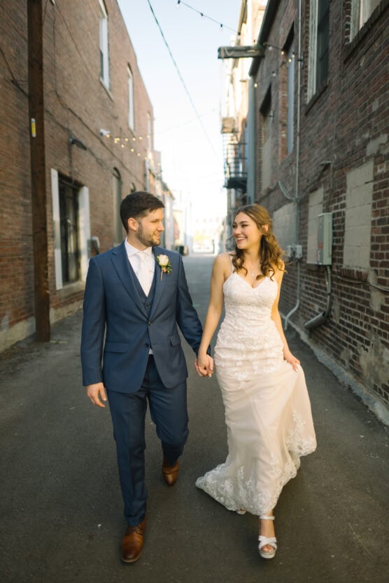 A bride and groom walk hand in hand down a sunlit alleyway, smiling at each other. The groom wears a blue suit; the bride wears a white lace gown. String lights and brick buildings line the alley. by Justin Salem Meyer