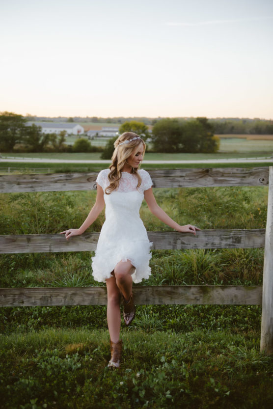 A woman in a short white lace dress and cowboy boots leans against a wooden fence. She stands in a grassy field with trees and farm buildings in the background at sunset. by Justin Salem Meyer