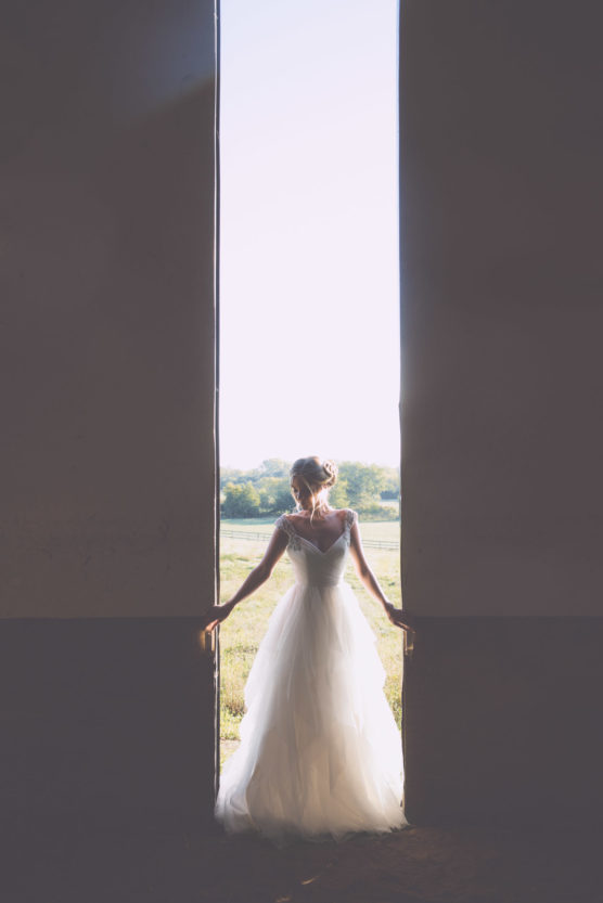 A bride in a white wedding dress stands in a large doorway, holding each side, with sunlight streaming in behind her and a scenic outdoor landscape visible beyond. by Justin Salem Meyer