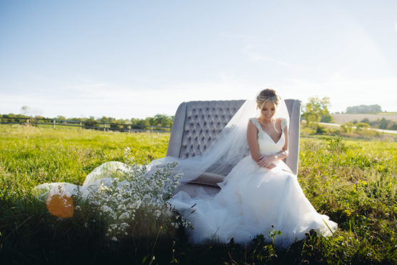A bride in a white gown and veil sits on an elegant bench in a sunny, grassy field, surrounded by white flowers, with rolling hills and blue sky in the background. by Justin Salem Meyer