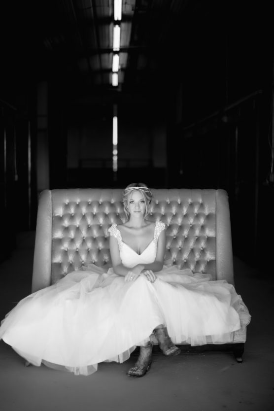 A woman in a wedding dress and boots sits on a tufted sofa, hands folded in her lap, looking at the camera. The background is dark, drawing focus to her in the softly lit scene. by Justin Salem Meyer