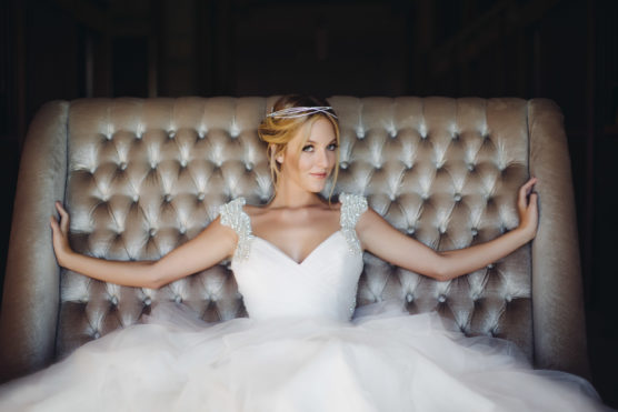 A woman in a white wedding dress with jeweled shoulder accents sits on a tufted, silver velvet couch, arms outstretched along the back. She wears a delicate headpiece and looks off to the side, smiling softly. by Justin Salem Meyer