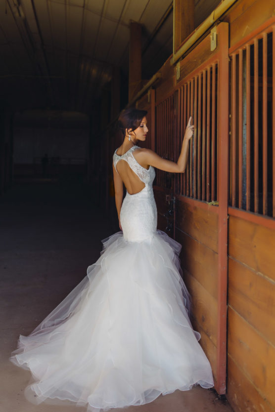A woman in a white, fitted, mermaid-style wedding dress with a tulle skirt stands indoors by a wooden stable door, looking to the side and touching the slats, with light streaming in from behind. by Justin Salem Meyer