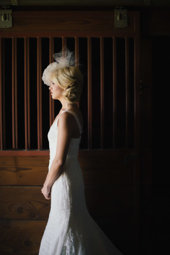 A woman in a white lace wedding dress and fascinator stands in profile against a dark wooden backdrop, softly lit from the side. by Justin Salem Meyer