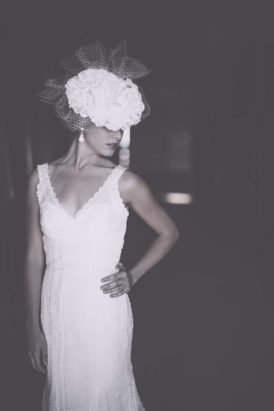 A woman in a sleeveless white lace dress poses with one hand on her hip, wearing a large, flowered headpiece with netting that partially covers her eyes, in a dimly lit setting. by Justin Salem Meyer