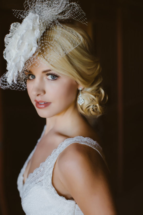 A woman in a white lace dress with blonde hair styled in loose curls, wearing a large white floral fascinator with netting and ornate drop earrings, looks at the camera with a soft, elegant expression. by Justin Salem Meyer