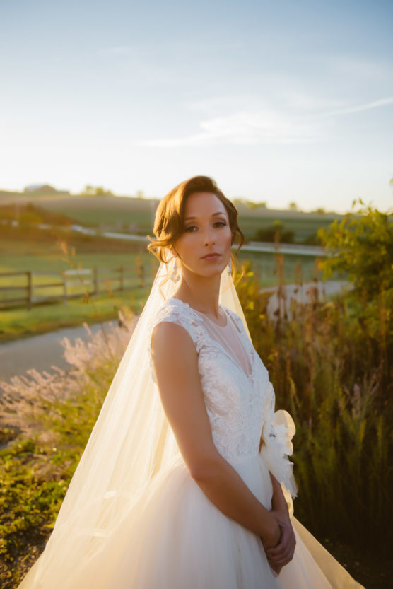 A bride in a white lace wedding dress and veil stands outdoors at sunset, surrounded by greenery and wildflowers, with a wooden fence and fields in the background. by Justin Salem Meyer