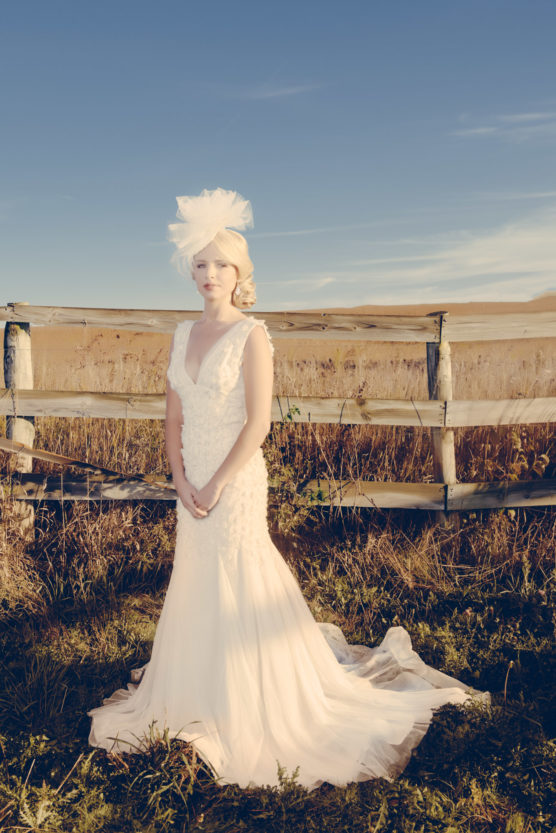 A woman in a white wedding dress and a decorative headpiece stands in front of a rustic wooden fence, outdoors in a sunlit field under a clear blue sky. by Justin Salem Meyer