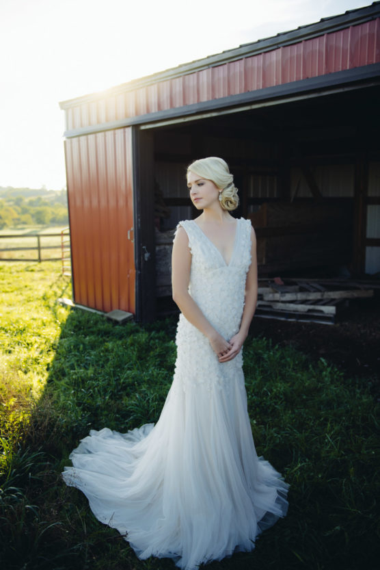 A woman in a white wedding dress stands in front of a red barn, looking to the side. Sunlight shines from behind, casting a soft glow over the grassy area and her gown. by Justin Salem Meyer