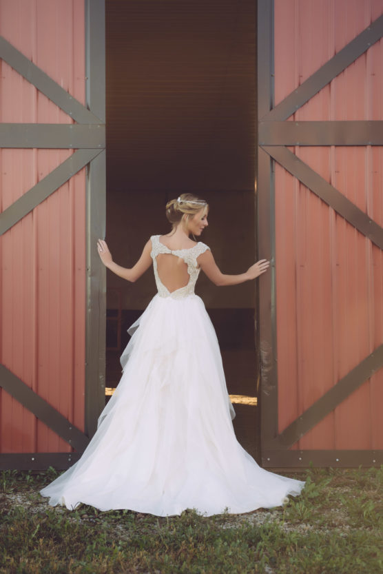 A woman in a white, backless wedding dress stands in an open barn doorway, facing away and holding each side of the door. The barn has large red and brown doors, and sunlight highlights her dress and hair. by Justin Salem Meyer