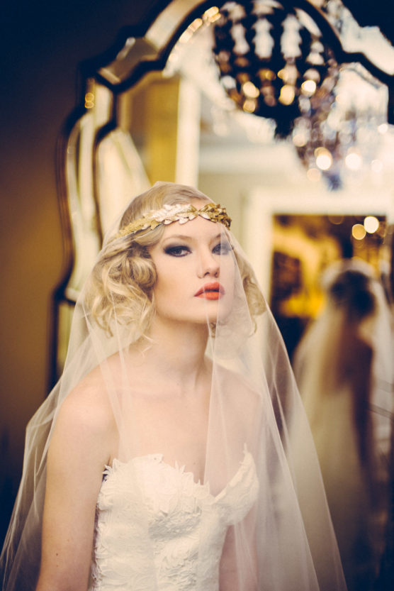 A bride with wavy blonde hair, wearing a strapless white wedding dress, a gold headpiece, and a sheer veil stands in front of an ornate mirror in soft, warm lighting. by Justin Salem Meyer
