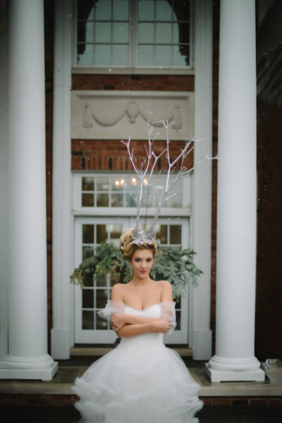 A woman in a white, airy dress with bare shoulders stands between two white columns. She wears a crown with silver branches, has her arms crossed, and stands in front of a large window and greenery. by Justin Salem Meyer
