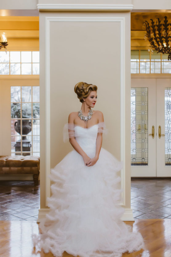 A woman in a strapless, layered white gown with tulle stands indoors against a cream-colored pillar. She wears an ornate necklace and has an elegant updo. Large glass doors and windows are visible in the background. by Justin Salem Meyer