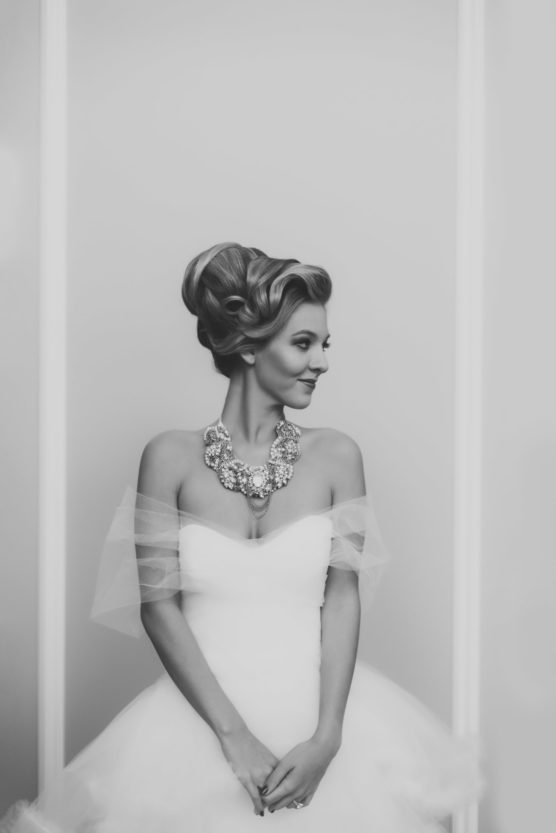 A woman in a strapless white gown with tulle sleeves stands with her hands clasped, wearing an ornate necklace and her hair styled in an elegant updo, gazing to the side against a plain background. by Justin Salem Meyer