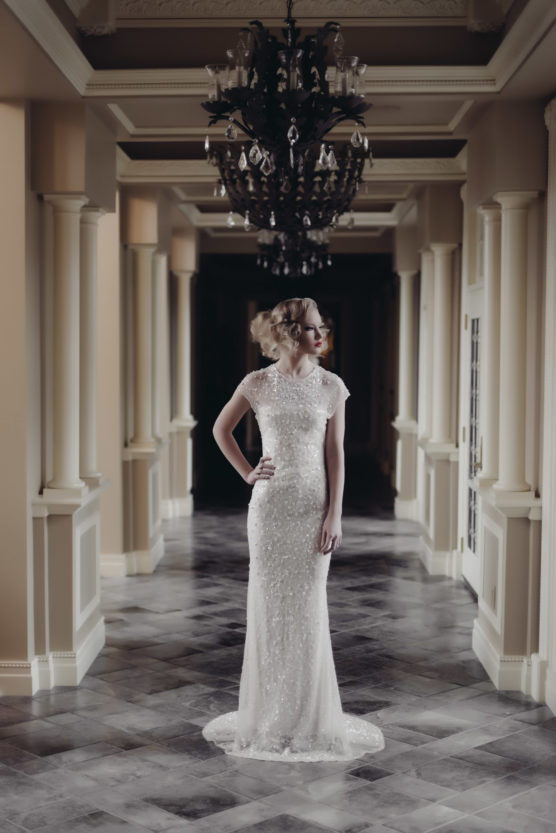 A woman in a fitted, elegant white gown stands in a grand hallway with high ceilings, ornate chandeliers, and columns. She gazes to the side, with one hand on her hip, her hair styled in soft waves. by Justin Salem Meyer