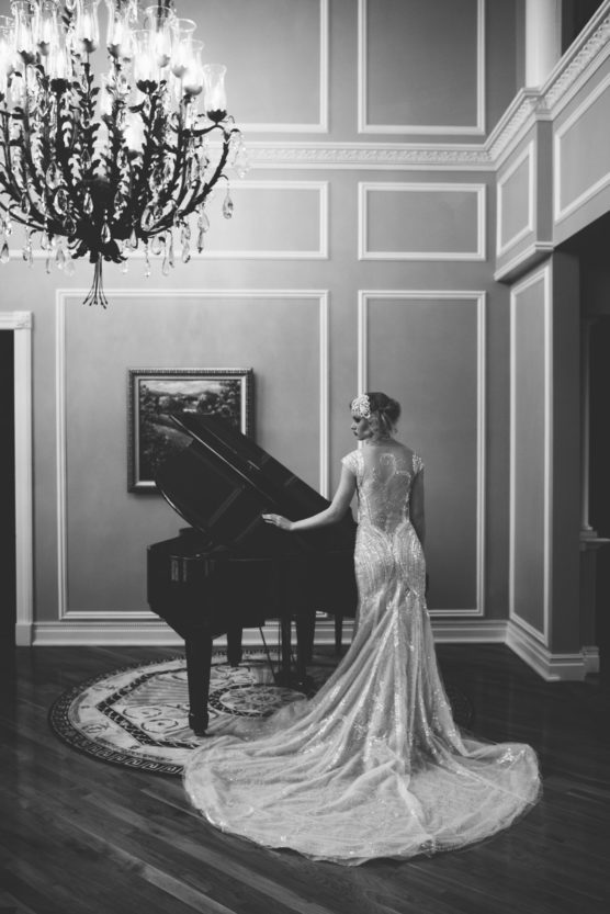 A woman in an elegant, long gown stands beside a grand piano in a sophisticated room with wooden floors, a chandelier, and framed artwork on the wall. The image is in black and white. by Justin Salem Meyer