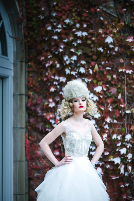 A woman in a white, sleeveless wedding dress and a large, fluffy fur hat stands with hands on hips in front of a wall covered in red and green ivy. by Justin Salem Meyer