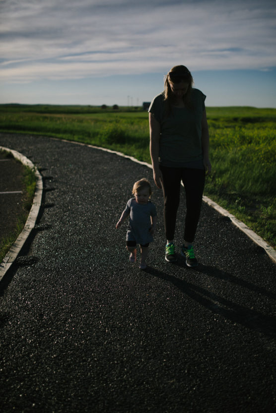 An adult and a small child walk together on a winding path through a grassy field under a partly cloudy sky. The adult holds the child’s hand, casting long shadows in the late afternoon light. by Justin Salem Meyer