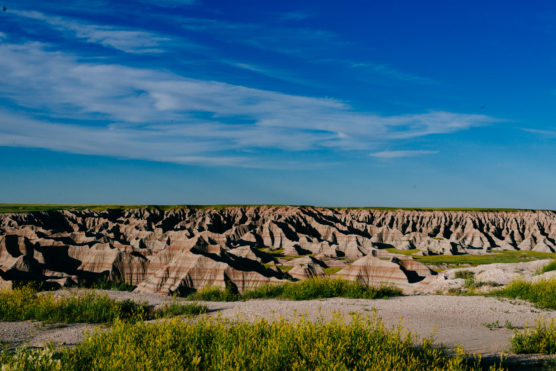 Eroded rock formations in a vast, rugged landscape under a blue sky with wispy clouds, framed by green grass and yellow wildflowers in the foreground. by Justin Salem Meyer
