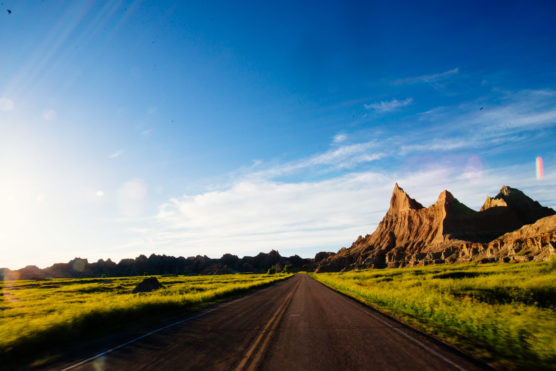 A paved road stretches toward jagged, sunlit rock formations under a clear blue sky, bordered by green grassy fields, with sunlight and lens flares on the left side. by Justin Salem Meyer