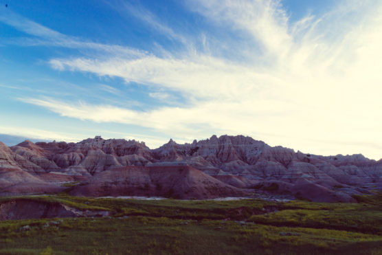 Eroded rock formations rise above green grass under a wide, blue sky with wispy white clouds, creating a dramatic landscape. by Justin Salem Meyer