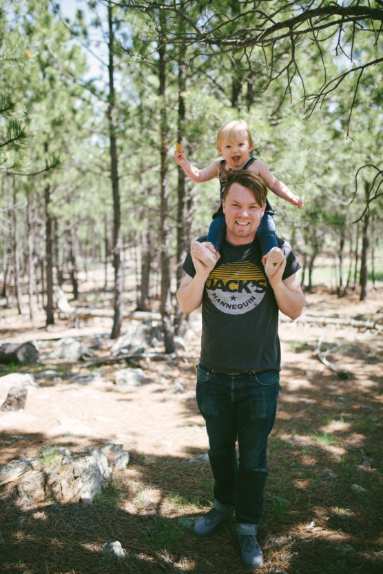 A man stands in a sunlit forest with a young child sitting on his shoulders. Both are smiling and holding hands, surrounded by tall trees and sunlight filtering through the branches. by Justin Salem Meyer