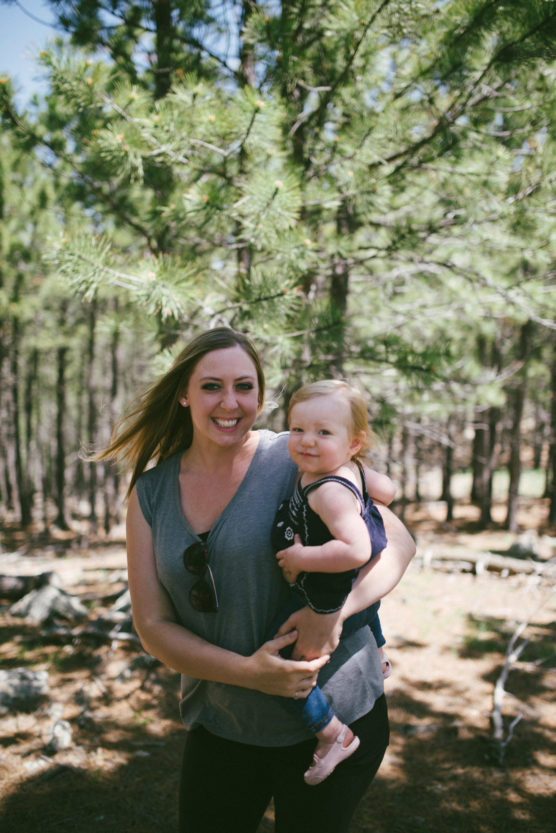 A woman smiles while holding a toddler in her arms outdoors. They are standing among tall pine trees with sunlight filtering through the branches. Both appear happy and relaxed in the natural setting. by Justin Salem Meyer