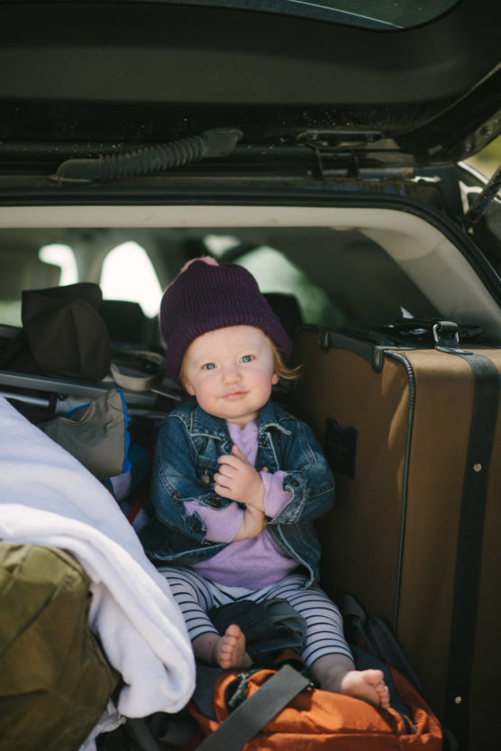 A young child wearing a purple beanie, denim jacket, and striped leggings sits barefoot among luggage in the back of a car, looking at the camera with a slight smile. by Justin Salem Meyer