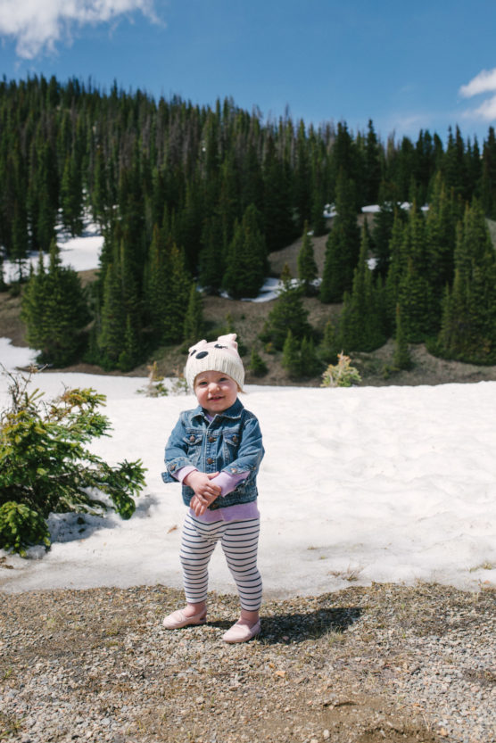 A small child wearing a white knit hat, denim jacket, and striped leggings stands and smiles on a snowy mountain landscape with evergreen trees and a partly cloudy blue sky in the background. by Justin Salem Meyer