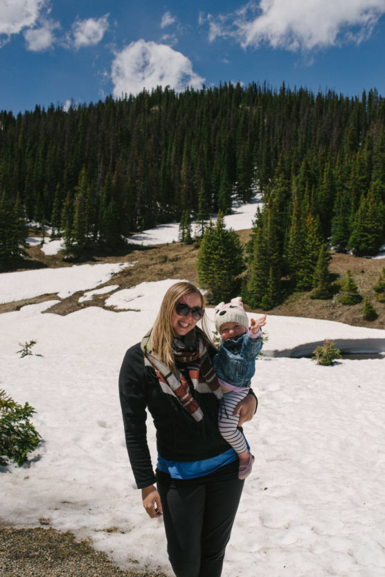A woman wearing sunglasses holds a baby in winter clothes on a snowy mountain landscape, with pine trees and a blue sky in the background. by Justin Salem Meyer