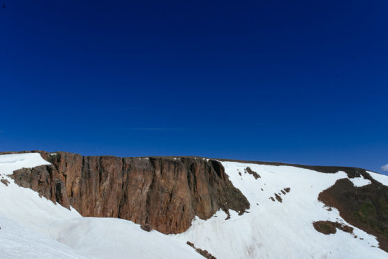 Cliffs of brown rock rise above snow-covered slopes under a deep blue, cloudless sky. The snow partially covers the ground, contrasting with the rugged rock face. by Justin Salem Meyer