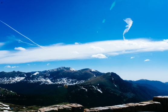 Snow-capped mountains under a bright blue sky with a few clouds and visible airplane contrails, viewed from a high vantage point with part of a stone wall in the foreground. by Justin Salem Meyer