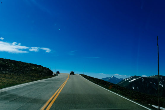 A mountain road with two cars driving uphill under a clear blue sky, with snow-capped peaks visible in the background and yellow lines dividing the asphalt. by Justin Salem Meyer