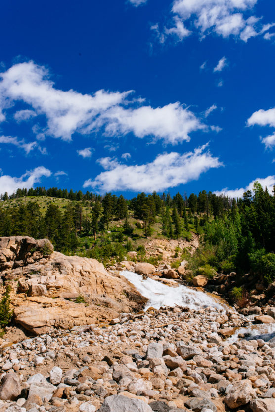 A rocky stream flows down a hillside with scattered boulders, surrounded by green trees beneath a bright blue sky with scattered white clouds. by Justin Salem Meyer