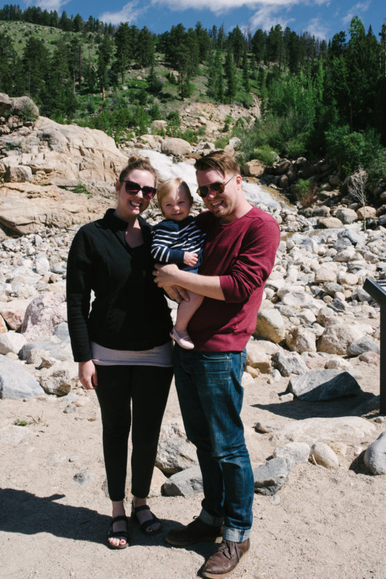 A smiling couple stands outdoors on rocky terrain, holding a small child between them. Pine trees and hills are visible in the background under a partly cloudy sky. by Justin Salem Meyer