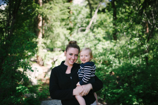 A woman with her hair in a bun, wearing a black jacket, holds a smiling baby dressed in a striped shirt. They stand together outdoors surrounded by greenery and trees. by Justin Salem Meyer