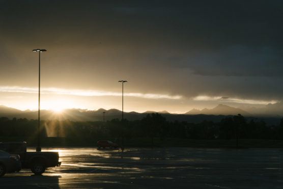 A wet, empty parking lot at sunset with mountains in the distance and dramatic clouds overhead. Sunlight breaks through the clouds, casting reflections on the pavement and illuminating two tall light poles. by Justin Salem Meyer