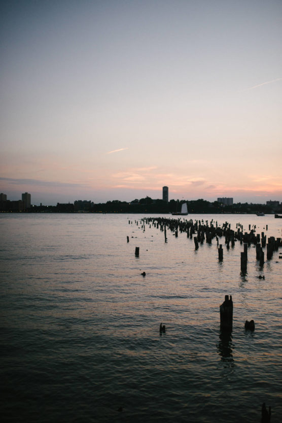 A calm body of water at sunset with scattered wooden posts emerging from the surface, city buildings silhouetted in the distance against a pastel sky. by Justin Salem Meyer
