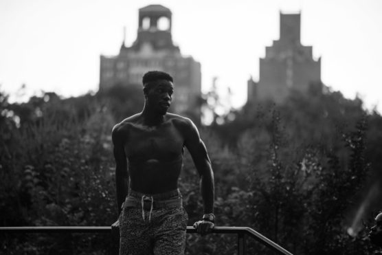 A shirtless man leans on a railing outdoors, with blurred tall buildings and trees in the background; the photo is in black and white, giving a dramatic and moody atmosphere. by Justin Salem Meyer