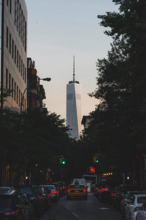 A city street scene at dusk shows cars stopped at a traffic light, with the tall One World Trade Center building towering in the background, framed by trees and surrounding buildings. by Justin Salem Meyer