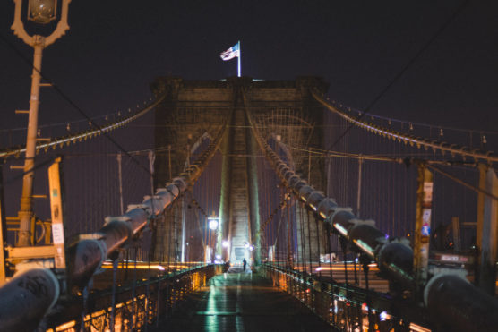 Night view of the Brooklyn Bridge in New York City, with cables illuminated and an American flag flying atop the stone towers, and blurred lights from passing vehicles. by Justin Salem Meyer