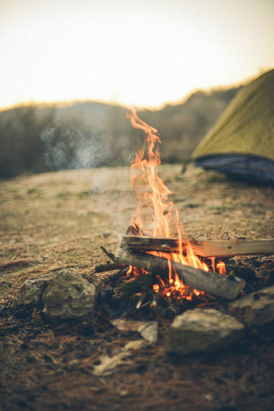 A close-up of a campfire burning with logs and surrounded by rocks, set outdoors on dry ground, with a yellow tent and blurred hills visible in the background. by Justin Salem Meyer